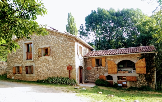A rustic stone house with wooden shutters and a red-tiled roof is set amid lush greenery. The building features multiple small windows with wooden frames and a signpost near the entrance. The setting suggests a peaceful, rural environment, potentially in the countryside.
