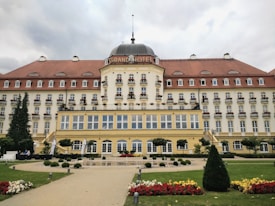 A grand, multi-story hotel building with a dome in the center, adorned with numerous windows and balconies. The structure features a red-tiled roof and a cream-colored facade. Manicured hedges and vibrant flowers line the pathway leading to the entrance, surrounded by a spacious lawn.