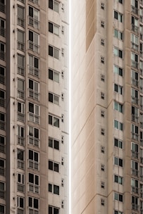 A narrow vertical gap between two tall apartment buildings. The structures have a modern architectural design with multiple windows visible, some with small balconies. The buildings have a uniform, off-white color, adding a sense of urban density and minimalism.