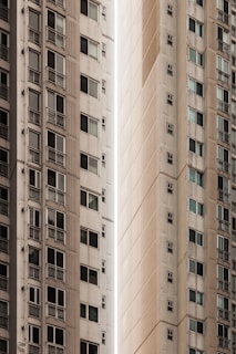 A narrow vertical gap between two tall apartment buildings. The structures have a modern architectural design with multiple windows visible, some with small balconies. The buildings have a uniform, off-white color, adding a sense of urban density and minimalism.