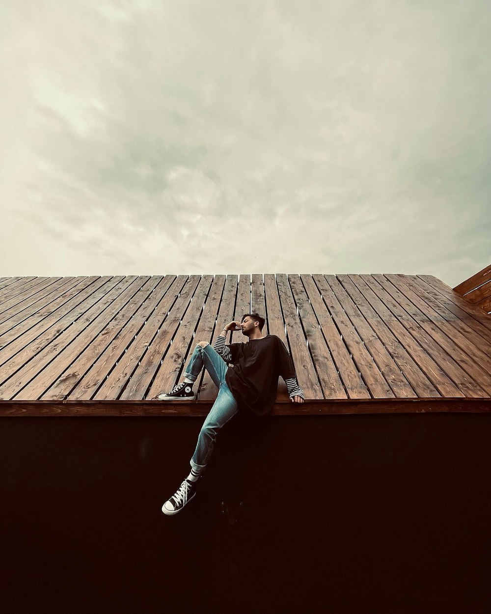 man in black jacket and blue denim jeans sitting on brown wooden roof