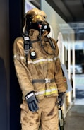A mannequin on display is wearing a full firefighter suit, which includes a helmet, gloves, and respiratory equipment. The suit appears to be used or designed to look worn, with visible signs of dirt and soot. The setting seems to be an indoor environment, possibly a shop or museum, with reflections on the glass indicating an outdoor scene.