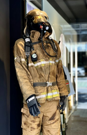 A mannequin on display is wearing a full firefighter suit, which includes a helmet, gloves, and respiratory equipment. The suit appears to be used or designed to look worn, with visible signs of dirt and soot. The setting seems to be an indoor environment, possibly a shop or museum, with reflections on the glass indicating an outdoor scene.