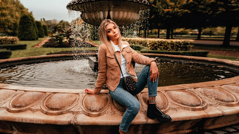 woman in brown blazer and blue denim jeans sitting on brown concrete fountain during daytime