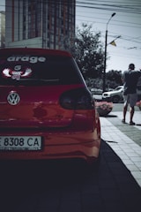A red Volkswagen car is parked on a street, with a prominent decal on the rear window. A person stands nearby, holding a phone and wearing a cap. Urban elements, such as a high-rise building, street lamp, and power lines, are visible in the background.