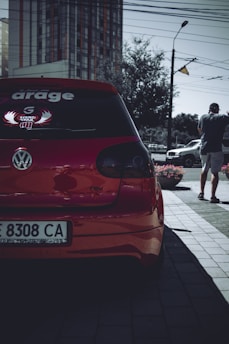 A red Volkswagen car is parked on a street, with a prominent decal on the rear window. A person stands nearby, holding a phone and wearing a cap. Urban elements, such as a high-rise building, street lamp, and power lines, are visible in the background.