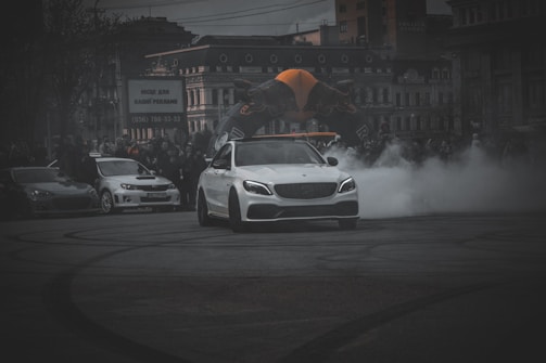 A white sports car is drifting on a street in front of a crowd of spectators, with smoke billowing from its tires. Several other cars are parked in the background. The event appears to be part of a car show or racing event in an urban area with buildings and an inflatable figure visible in the distance.