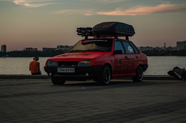 A red car with 'Street Sound' decals is parked near a waterfront during sunset. The car has a roof box and is situated on a paved area. In the background, a person in an orange shirt sits by the water, and city buildings are visible across the water.