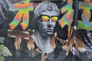 A colorful graffiti mural featuring a classical-style bust wearing vibrant, reflective sunglasses. Surrounding the face, there are bold, psychedelic patterns with bright neon colors. The art blends traditional sculpture aesthetics with modern street art techniques.