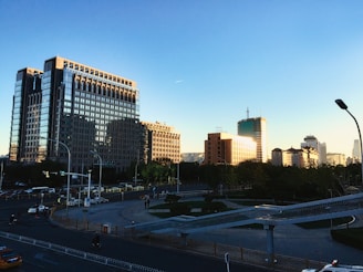 A cityscape featuring modern high-rise buildings with reflective glass exteriors under a clear blue sky. The structures are bathed in warm sunlight, creating a contrasting light effect. Below, a street with sparse traffic and a pedestrian area with walkways are visible, accompanied by a few scattered vehicles and streetlights.
