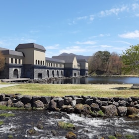 A building with a modern, industrial design sits beside a calm lake. The structure is constructed with light-colored walls and dark roofs, featuring numerous arches. In the foreground, a small stream flows over rocks with green grass surrounding it. Sparse trees standing nearby hint at a natural scenic background blending into blue skies.