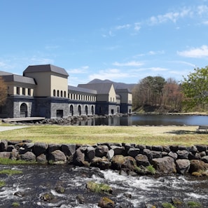 A building with a modern, industrial design sits beside a calm lake. The structure is constructed with light-colored walls and dark roofs, featuring numerous arches. In the foreground, a small stream flows over rocks with green grass surrounding it. Sparse trees standing nearby hint at a natural scenic background blending into blue skies.