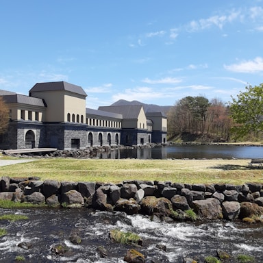 A building with a modern, industrial design sits beside a calm lake. The structure is constructed with light-colored walls and dark roofs, featuring numerous arches. In the foreground, a small stream flows over rocks with green grass surrounding it. Sparse trees standing nearby hint at a natural scenic background blending into blue skies.