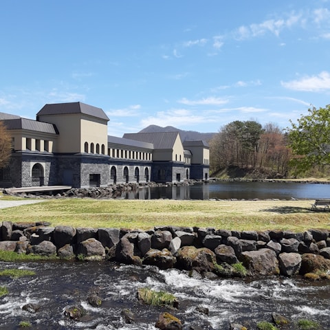 A building with a modern, industrial design sits beside a calm lake. The structure is constructed with light-colored walls and dark roofs, featuring numerous arches. In the foreground, a small stream flows over rocks with green grass surrounding it. Sparse trees standing nearby hint at a natural scenic background blending into blue skies.