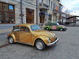A vintage Volkswagen Beetle covered in a shimmering gold exterior is parked on a cobblestone street. In the background, another Beetle with a vibrant, artistic paint job featuring a large rose is visible. The scene is set in an urban environment with classic European architecture, including storefronts and flags hanging from buildings.