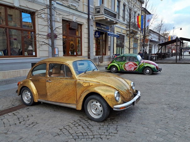 A vintage Volkswagen Beetle covered in a shimmering gold exterior is parked on a cobblestone street. In the background, another Beetle with a vibrant, artistic paint job featuring a large rose is visible. The scene is set in an urban environment with classic European architecture, including storefronts and flags hanging from buildings.