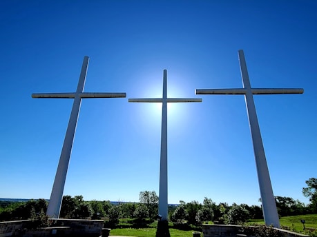 Three large crosses are silhouetted against a clear blue sky with the sun positioned directly behind the central cross. The crosses are set on an elevated platform, surrounded by greenery including trees and bushes.