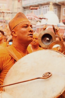 A man is holding and playing a large traditional drum amidst a crowd in what appears to be a lively procession or festival. The background includes other participants, and ornate buildings, with some people holding instruments.