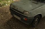 A worn and weathered small car, possibly from the late 1980s or early 1990s. The paint is faded and discolored, with visible rust and grime. The headlight covers are slightly yellowed. The scene is dimly lit, suggesting it might be taken in low light or shaded area.