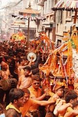 A lively street scene featuring a large group of people participating in a traditional festival procession. Many participants are wearing orange shirts and are carrying elaborately decorated structures adorned with marigold garlands and colorful parasols. Musicians are playing drums and other instruments, contributing to the vibrant atmosphere. Buildings line the narrow street, enhancing the festive ambiance.