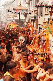 A lively street scene featuring a large group of people participating in a traditional festival procession. Many participants are wearing orange shirts and are carrying elaborately decorated structures adorned with marigold garlands and colorful parasols. Musicians are playing drums and other instruments, contributing to the vibrant atmosphere. Buildings line the narrow street, enhancing the festive ambiance.