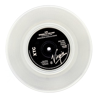 A clear vinyl record with a black label in the center, displaying the text 'Life Begins At The Hop' by XTC, produced by Steve Lillywhite. The label features the Virgin logo and various technical details about the record.