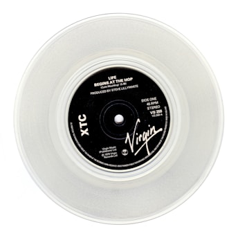 A clear vinyl record with a black label in the center, displaying the text 'Life Begins At The Hop' by XTC, produced by Steve Lillywhite. The label features the Virgin logo and various technical details about the record.