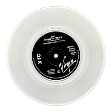 A clear vinyl record with a black label in the center, displaying the text 'Life Begins At The Hop' by XTC, produced by Steve Lillywhite. The label features the Virgin logo and various technical details about the record.