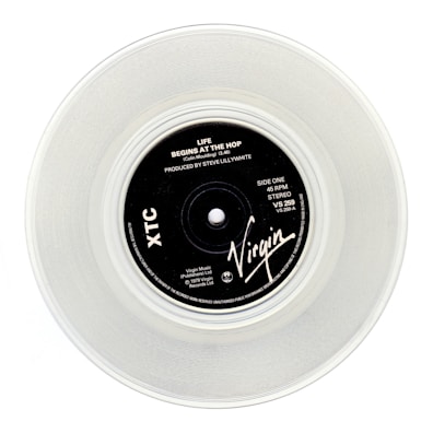 A clear vinyl record with a black label in the center, displaying the text 'Life Begins At The Hop' by XTC, produced by Steve Lillywhite. The label features the Virgin logo and various technical details about the record.