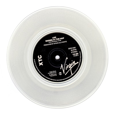 A clear vinyl record with a black label in the center, displaying the text 'Life Begins At The Hop' by XTC, produced by Steve Lillywhite. The label features the Virgin logo and various technical details about the record.