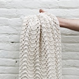 A hand holding a cream-colored knitted blanket with a textured pattern against a white brick wall.