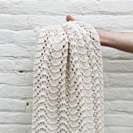 A hand holding a cream-colored knitted blanket with a textured pattern against a white brick wall.