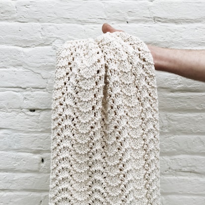 A hand holding a cream-colored knitted blanket with a textured pattern against a white brick wall.