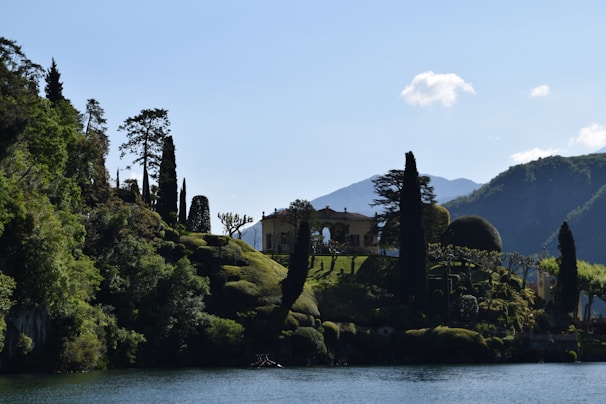 A picturesque scene featuring a villa surrounded by lush greenery on a hillside. Tall trees and manicured gardens decorate the landscape, set against a backdrop of mountains. The foreground includes a body of water reflecting the natural beauty of the surroundings.