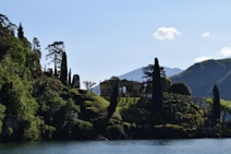 A picturesque scene featuring a villa surrounded by lush greenery on a hillside. Tall trees and manicured gardens decorate the landscape, set against a backdrop of mountains. The foreground includes a body of water reflecting the natural beauty of the surroundings.