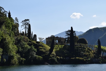 A picturesque scene featuring a villa surrounded by lush greenery on a hillside. Tall trees and manicured gardens decorate the landscape, set against a backdrop of mountains. The foreground includes a body of water reflecting the natural beauty of the surroundings.