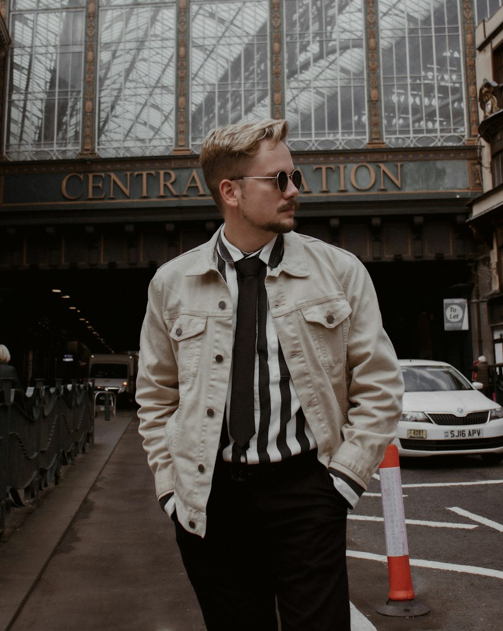 man in beige button up jacket and black pants standing on sidewalk during daytime