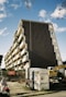 A modern apartment building with a distinctive geometric design, featuring several balconies. Below, there is construction equipment and a fenced-off area, indicating ongoing construction or renovation work. A woman with a shopping trolley is walking past on the sidewalk. The sky is partly cloudy.
