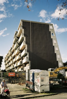 A modern apartment building with a distinctive geometric design, featuring several balconies. Below, there is construction equipment and a fenced-off area, indicating ongoing construction or renovation work. A woman with a shopping trolley is walking past on the sidewalk. The sky is partly cloudy.