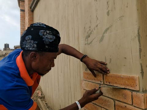 A person wearing a bandana and a blue and orange shirt is working on a brick wall, carefully placing a brick into place. The wall shows signs of cement application, and there is construction material scattered around. A building and a water tank are visible in the background.