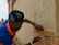 A person wearing a bandana and a blue and orange shirt is working on a brick wall, carefully placing a brick into place. The wall shows signs of cement application, and there is construction material scattered around. A building and a water tank are visible in the background.