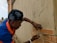 A person wearing a bandana and a blue and orange shirt is working on a brick wall, carefully placing a brick into place. The wall shows signs of cement application, and there is construction material scattered around. A building and a water tank are visible in the background.