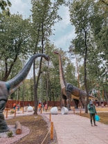 Life-sized dinosaur sculptures are situated along a wooden path in a park with tall trees. People are walking around, some carrying bags and wearing colorful jackets. Large replica dinosaur eggs are also placed on the ground.
