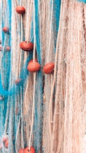 A close-up view of a tangled mass of fishing nets featuring vibrant blue and white threads interwoven with thick ropes. The nets are adorned with prominent red-brown floats spaced along the ropes, creating a textured and intricate pattern.