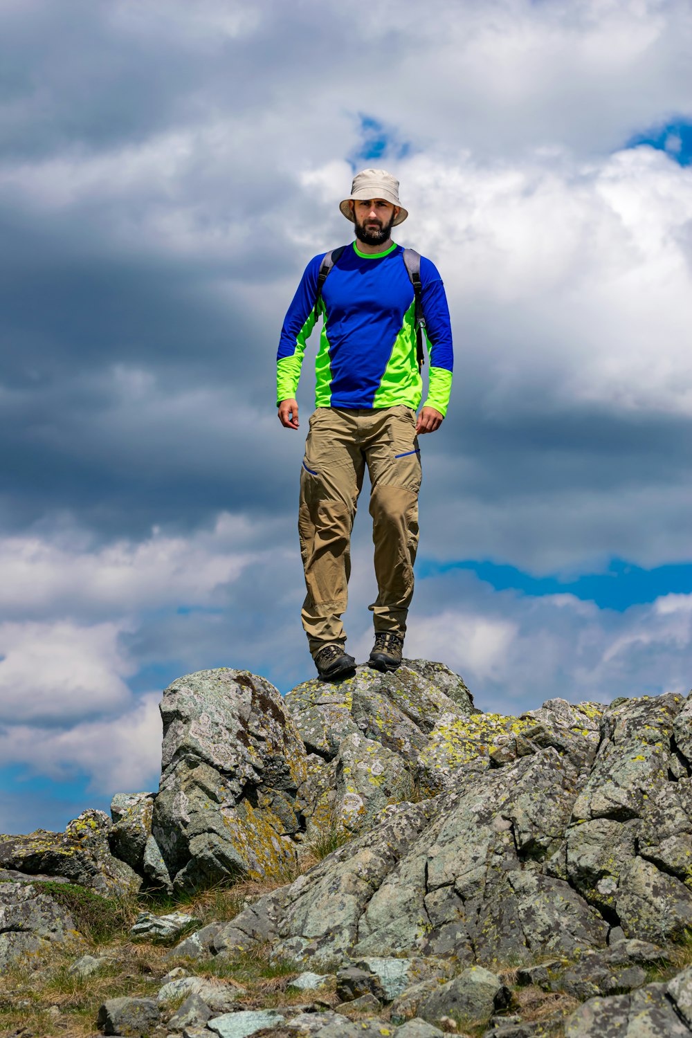 man in blue jacket and brown pants standing on rock under white clouds during daytime
