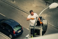A man is working on a streetlight, standing on a hydraulic lift on a city street. He is wearing a white shirt and orange gloves. Below, a blue car is parked on the road, with visible tire tracks and road markings. The pavement features a grid pattern with cobblestones.
