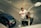 A man is working on a streetlight, standing on a hydraulic lift on a city street. He is wearing a white shirt and orange gloves. Below, a blue car is parked on the road, with visible tire tracks and road markings. The pavement features a grid pattern with cobblestones.