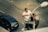 A man is working on a streetlight, standing on a hydraulic lift on a city street. He is wearing a white shirt and orange gloves. Below, a blue car is parked on the road, with visible tire tracks and road markings. The pavement features a grid pattern with cobblestones.