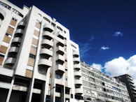 A view of a residential building with a modern architectural design featuring rounded balconies. The sky is bright blue with some white clouds, creating a vibrant contrast. The building is mostly white with sections of tan-colored wall panels.