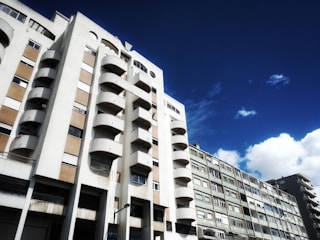 A view of a residential building with a modern architectural design featuring rounded balconies. The sky is bright blue with some white clouds, creating a vibrant contrast. The building is mostly white with sections of tan-colored wall panels.